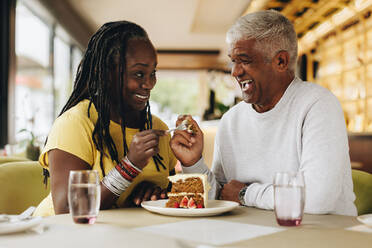 Ein älteres Paar lächelt sich gegenseitig an, während es einen leckeren Kuchen in einem Café isst. Ein glückliches älteres Paar, das sich in einem Restaurant amüsiert. Ein fröhliches reifes Paar, das seinen Ruhestand zusammen genießt. - JLPSF10469