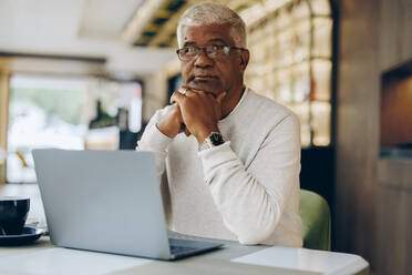 Älterer Geschäftsmann, der in die Kamera schaut, während er in einem modernen Café arbeitet. Älterer Geschäftsmann, der einen Laptop benutzt, während er aus der Ferne arbeitet. - JLPSF10488