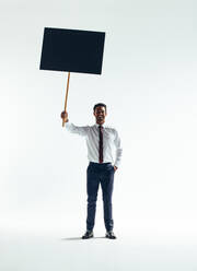 Glücklicher Geschäftsmann, der in die Kamera lächelt, während er ein Schild hochhält. Erfolgreicher Geschäftsmann, der eine Botschaft verbreitet, während er vor einem weißen Hintergrund steht. - JLPSF10528