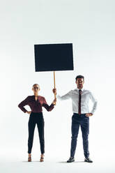 Zwei seriöse Geschäftsleute schauen in die Kamera, während sie ein schwarzes Plakat in einem Studio hochhalten. Zwei junge Geschäftsleute protestieren für die Gleichstellung der Geschlechter am Arbeitsplatz. - JLPSF10535