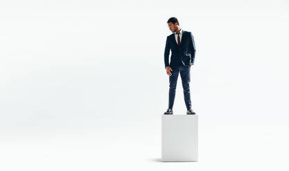 Geschäftsmann, der auf seine Hindernisse hinunterblickt, während er an der Spitze steht. Erfolgreicher Geschäftsmann, der auf einem hohen Block vor einem weißen Hintergrund steht. - JLPSF10569