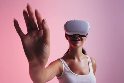 Fröhliche junge Frau, die mit ihrer Hand im virtuellen Raum interagiert, während sie eine Virtual-Reality-Brille trägt. Glückliche junge Frau, die eine 3D-Simulation in einem Studio erlebt. - JLPSF10624