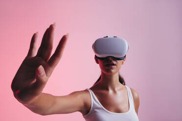 Junge Frau, die mit ihrer Hand im virtuellen Raum interagiert, während sie ein Virtual-Reality-Headset trägt. Weibliche Spielerin, die eine 3D-Simulation in einem Studio erlebt. - JLPSF10625