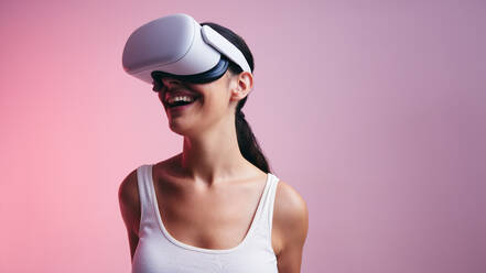 Cheerful young woman entering a fun virtual reality game in a studio. Woman laughing happily while wearing a virtual reality headset. Female gamer exploring 3D technology. - JLPSF10629