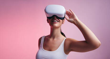 Happy young woman smiling cheerfully while wearing virtual reality goggles. Woman entering a fun virtual reality game. Female gamer exploring 3D technology in a studio. - JLPSF10630