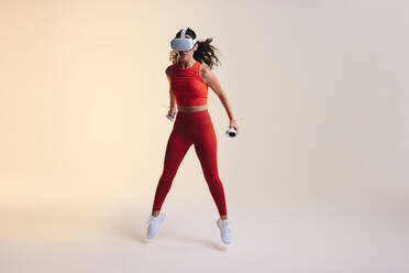 Sportlerin, die eine Trainingseinheit in der virtuellen Realität absolviert. Sportliche junge Frau, die in der Luft springt, während sie Gaming-Controller hält. Aktive junge Frau, die eine Virtual-Reality-Brille in einem Studio trägt. - JLPSF10644