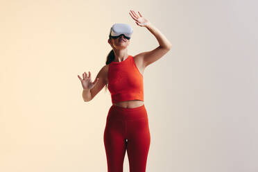 Glückliche junge Frau, die in einem Studio mit dem Metaversum interagiert. Aktive junge Frau, die sich umschaut, während sie ein Virtual-Reality-Headset trägt. Fröhliche junge Frau, die ein 3D-Spiel betritt. - JLPSF10678