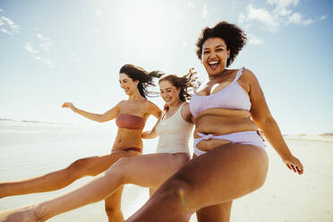 Three happy girlfriends having a good time at the beach. Group of playful female friends standing on one leg while wearing swimwear. Carefree young women laughing and enjoying their summer vacation. - JLPSF10734