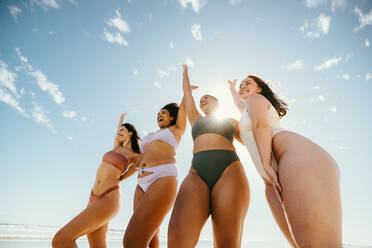 Eine Gruppe glücklicher Freundinnen, die mit erhobenen Armen jubeln, während sie Badekleidung am Strand tragen. Sorglose junge Frauen, die Spaß haben und ihren Urlaub genießen. - JLPSF10736