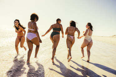 Bikini-Spaß. Gruppe unbeschwerter junger Frauen, die fröhlich lachen, während sie in Badekleidung am Strand laufen. Unbeschwerte Freundinnen, die während ihres Sommerurlaubs glückliche Erinnerungen schaffen. - JLPSF10747