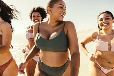 Frauen, die in Bikinis am Strand laufen. Eine Gruppe unbeschwerter junger Frauen, die fröhlich lächelnd in der Sonne laufen. Glückliche Freundinnen, die Spaß haben und ihren Sommerurlaub genießen. - JLPSF10749