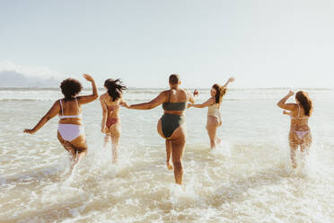 Spaß in Badekleidung. Rückansicht einer Gruppe von fröhlichen Freundinnen, die am Strand zusammen laufen und im Wasser planschen. Gruppe von unbeschwerten jungen Frauen, die Spaß haben und ihren Sommerurlaub genießen. - JLPSF10757
