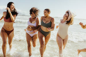 Fröhliche Freundinnen spritzen mit Wasser, während sie gemeinsam am Strand rennen. Eine Gruppe unbeschwerter junger Frauen, die ihren Sommerurlaub in Badekleidung genießen. - JLPSF10758