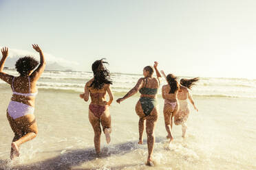 Rückansicht von glücklichen Freundinnen, die am Strand auf das Wasser zulaufen. Eine Gruppe unbeschwerter junger Frauen, die ihren Sommerurlaub in Badekleidung genießen. - JLPSF10759