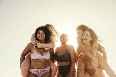 Freundinnen, die sich am Strand gegenseitig Huckepack nehmen. Eine Gruppe glücklicher junger Frauen, die fröhlich lächelnd in der Sommersonne stehen. Beste Freundinnen, die Spaß haben und ihren Urlaub gemeinsam genießen. - JLPSF10761