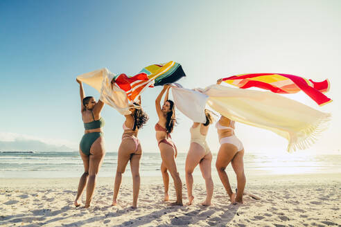 Frauen lassen am Strand ihre Handtücher fliegen. Rückansicht einer Gruppe glücklicher junger Frauen, die mit erhobenen Armen jubelnd beieinander stehen. Freundinnen, die in den Sommerferien Spaß haben. - JLPSF10764