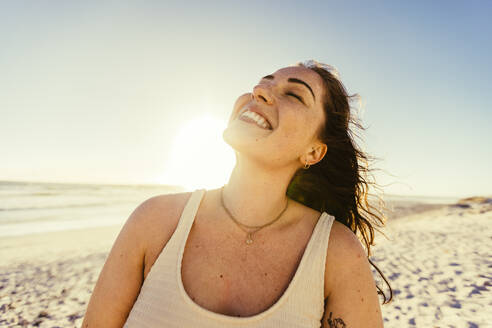 Embracing beach happiness. Cheerful young woman smiling with her eyes closed while standing at the beach at sunset. Carefree young woman enjoying her summer vacation in swimwear. - JLPSF10768