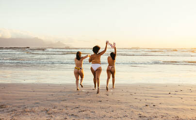 Frauen, die den Sommer in ihren natürlichen Körpern genießen. Rückansicht von drei barbusigen jungen Frauen, die in Bikinihöschen auf das Meerwasser zulaufen. Unbeschwerte Freundinnen, die im Urlaub Spaß haben. - JLPSF10781