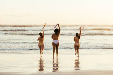 Drei glückliche junge Frauen, die in Bikinihöschen zum Wasser laufen. Eine Gruppe unbeschwerter Freundinnen hat Spaß und genießt ihren Urlaub am Strand. - JLPSF10782