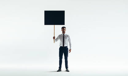 Selbstbewusster junger Geschäftsmann, der ein Plakat hochhält, während er vor einem weißen Hintergrund steht. Geschäftsaktivist, der in einem Studio für die Rechte der Arbeitnehmer protestiert. - JLPSF10808