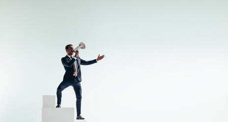 Successful businessman shouting with a megaphone while standing at the stop of a staircase. Cheerful businessman celebrating his victory after reaching his goal. - JLPSF10821