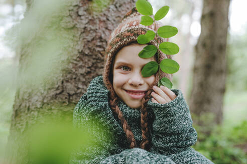 Fröhliches Mädchen mit Strickmütze sitzt an einem Baum im Wald - YTF00227