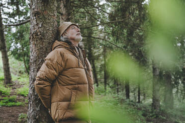 Senior man wearing jacket standing by tree in forest - YTF00237