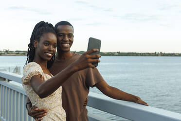Lächelndes junges Paar nimmt Selfie stehend am Fluss - JBUF00023