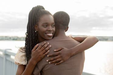Glückliche junge Frau umarmt Mann am Fluss bei Sonnenuntergang - JBUF00028