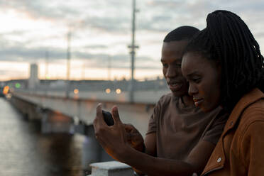 Junges Paar teilt sich ein Smartphone auf einer Brücke - JBUF00039