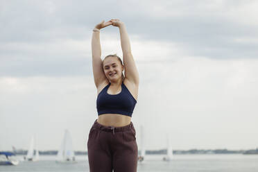 Smiling young woman doing stretching exercise in front of sea - JBUF00051