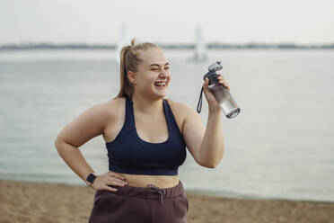 Happy young woman standing with water bottle on beach - JBUF00053