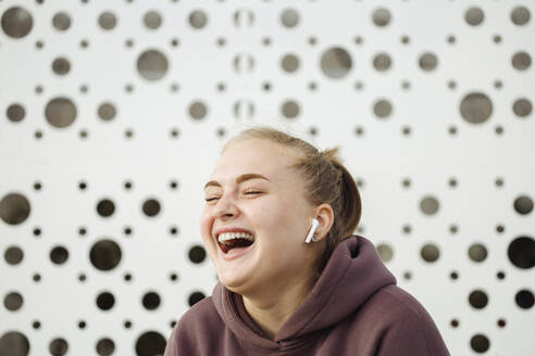 Fröhliche junge Frau mit drahtlosen In-Ear-Kopfhörern vor einer Wand - JBUF00056