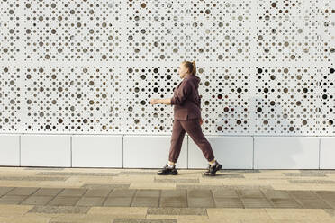 Overweight woman in hooded shirt walking along wall - JBUF00059