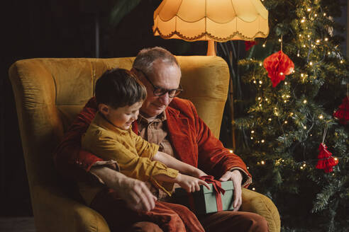 Glücklicher Enkel und Großvater mit einem Geschenkpaket zu Hause sitzend - VSNF00026