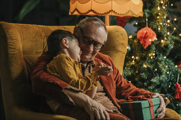 Happy grandfather and boy sitting with gift box by Christmas tree at home - VSNF00028