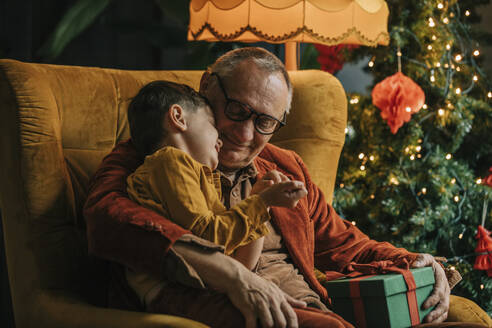 Glücklicher Großvater und Junge sitzen mit Geschenkpackung am Weihnachtsbaum zu Hause - VSNF00028