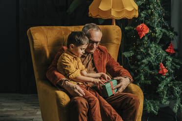 Grandson and grandfather sitting with gift box in armchair at home - VSNF00029