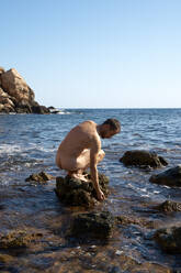 Nackter Mann sitzt auf einem Felsen inmitten des Meeres an einem sonnigen Tag - VEGF06057
