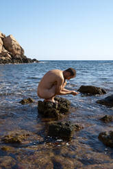 Nackter Mann in hockender Position auf einem Felsen am Meer - VEGF06058