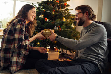 Ein Mann beschenkt seine Frau zu Hause, ein Paar sitzt am Weihnachtsbaum, tauscht Geschenke aus und lächelt. - JLPSF10868