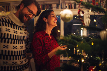 Mann und Frau lächelnd beim Schmücken des Weihnachtsbaums zu Hause. Ein Paar genießt die Vorbereitungen für den Weihnachtsabend zu Hause. - JLPSF10879