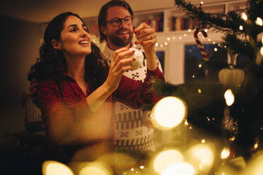 Ehepaar schmückt Weihnachtsbaum bei Familie zu Hause. Frau bindet glänzende dekorative Kugel auf Weihnachtsbaum mit Mann steht. - JLPSF10880