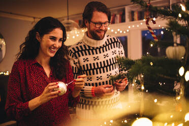 Eine Frau bindet eine glänzende dekorative Kugel an den Weihnachtsbaum, während ein Mann daneben steht. Ein Paar schmückt den Weihnachtsbaum bei einer Familie. - JLPSF10881