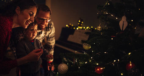 Eltern mit Sohn schmücken Weihnachtsbaum zu Hause. Junge Frau im Gespräch mit ihrem Sohn beim Schmücken des Weihnachtsbaums. - JLPSF10884