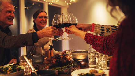 Familie stößt beim Weihnachtsessen mit Wein an. Die Familie genießt das gemeinsame Weihnachtsessen zu Hause, mit Fokus auf Hände und Weingläser. - JLPSF10889