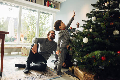 Ein Kind, das am Weihnachtsbaum steht und seine Hand ausstreckt, während die Eltern zusehen. Eine kleine Familie, die zu Weihnachten zu Hause zusammen ist. - JLPSF10901