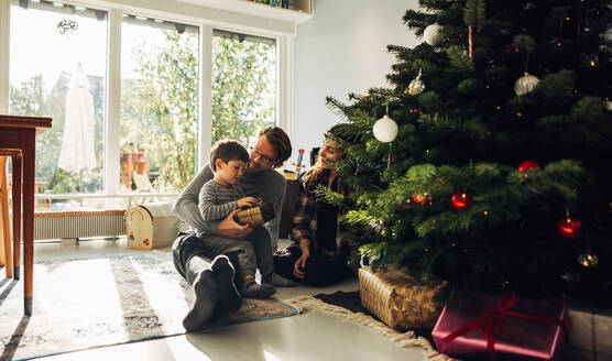 Familie feiert Weihnachten mit Geschenken zu Hause. Junge mit Geschenk in der Hand sitzt mit seinen Eltern am Weihnachtsbaum. - JLPSF10904