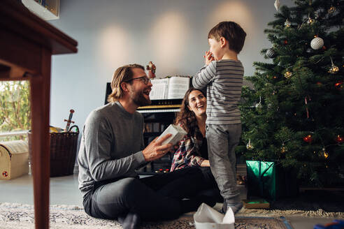 Eltern, die ihrem Sohn ein Geschenk überreichen, sitzen neben einem Weihnachtsbaum. Der Junge sieht aufgeregt aus, während er ein Weihnachtsgeschenk erhält. - JLPSF10908