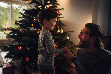 Man sitting by Christmas tree having fun with his son at home. Father and son enjoying at home during Christmas eve. - JLPSF10911